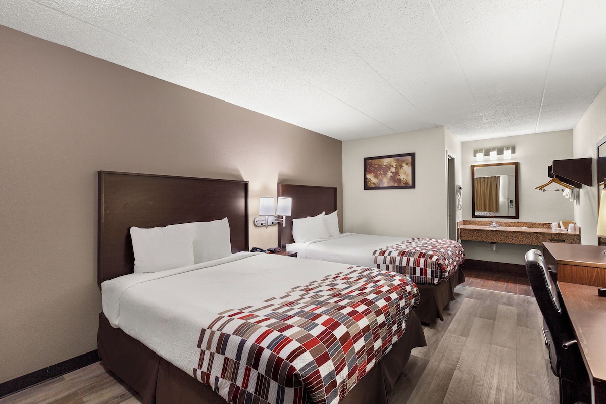 A hotel room with two beds, a desk, a chair, a mirror, and a bathroom with a sink and towel rack.