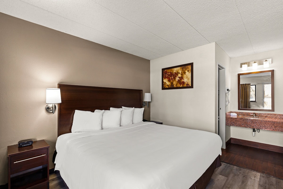 A hotel room with a large bed, a nightstand with a lamp, a painting on the wall, and a bathroom with a sink and mirror.