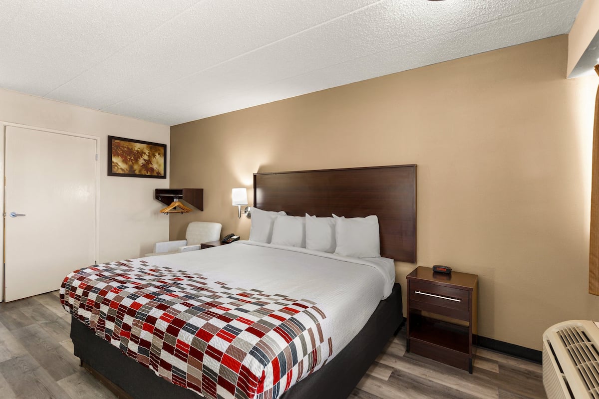 A hotel room with a large bed, a bedside table, a chair, a desk, and a wall-mounted lamp.