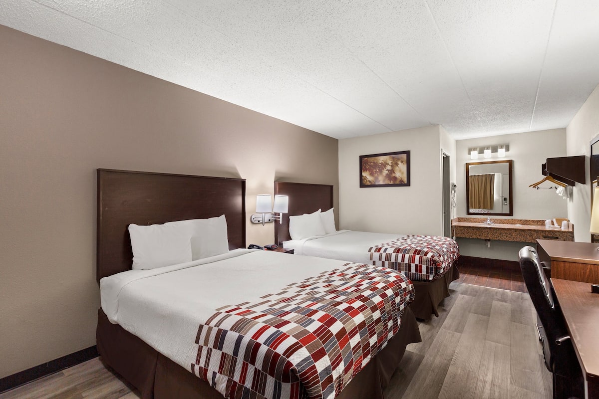 A hotel room with two beds, a desk, a chair, a mirror, and a bathroom with a sink and towel rack.