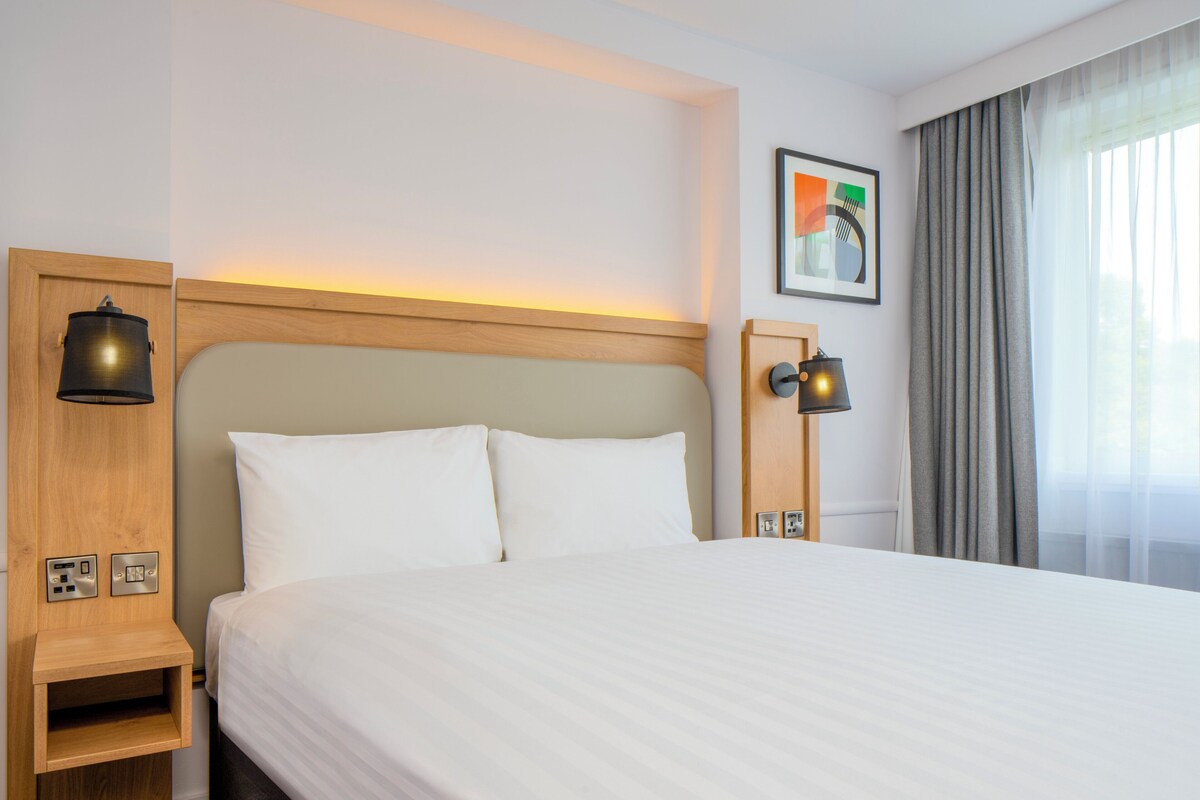 A hotel room with a large bed, wooden headboard, bedside lamps, a power outlet, and a window with curtains.