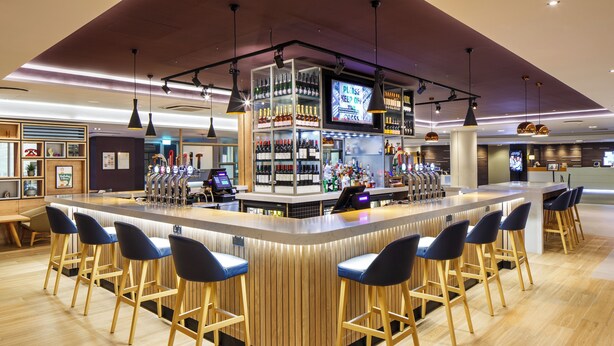 A bar area with a wooden counter, bar stools, and a television screen. - Holiday Inn London - Heathrow M4,jct.4 by IHG (West Drayton)