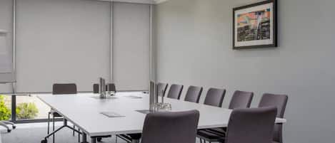 Un salón de conferencias con una larga mesa, sillas, una pantalla de proyector y un cuadro en la pared.
