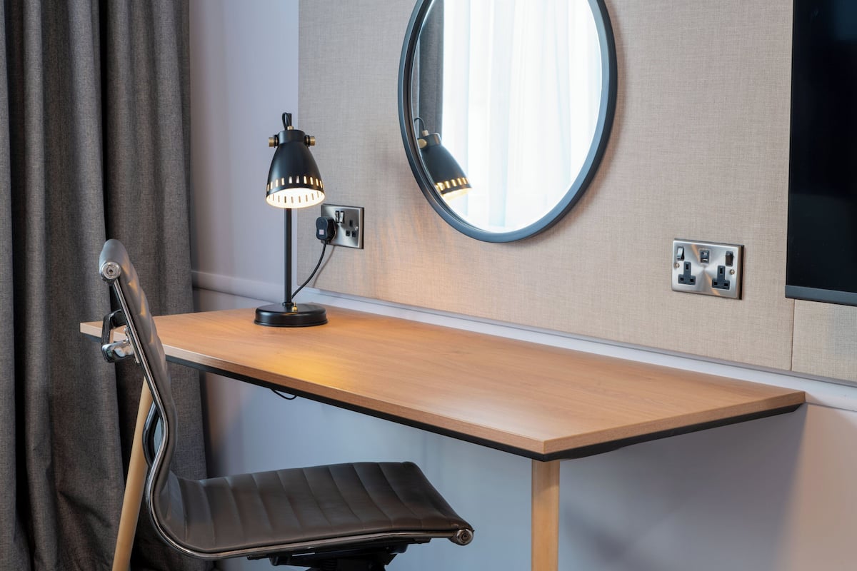 A desk with a lamp, a chair, a round mirror, and a power outlet.