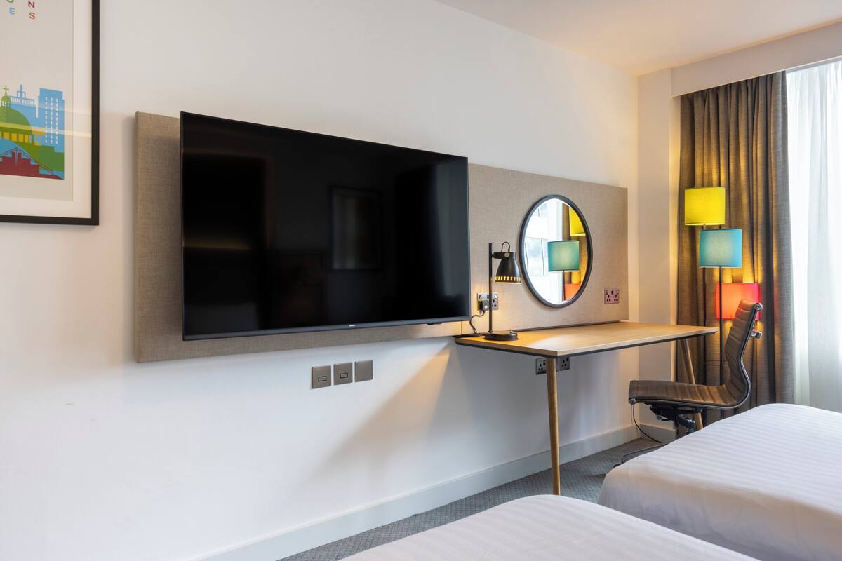 A hotel room with a flat-screen TV, a desk with a chair, a round mirror, and two beds.