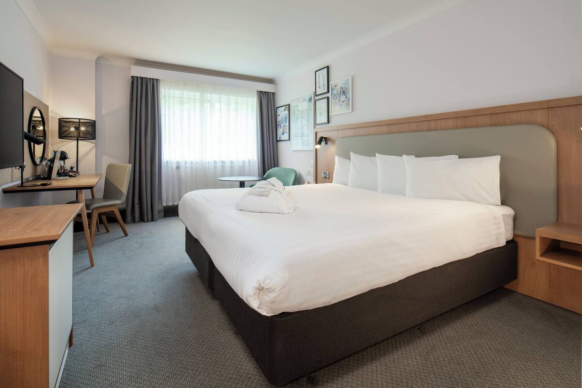 Une chambre d’hôtel avec un grand lit, un bureau, une chaise, une lampe et une fenêtre avec des rideaux.