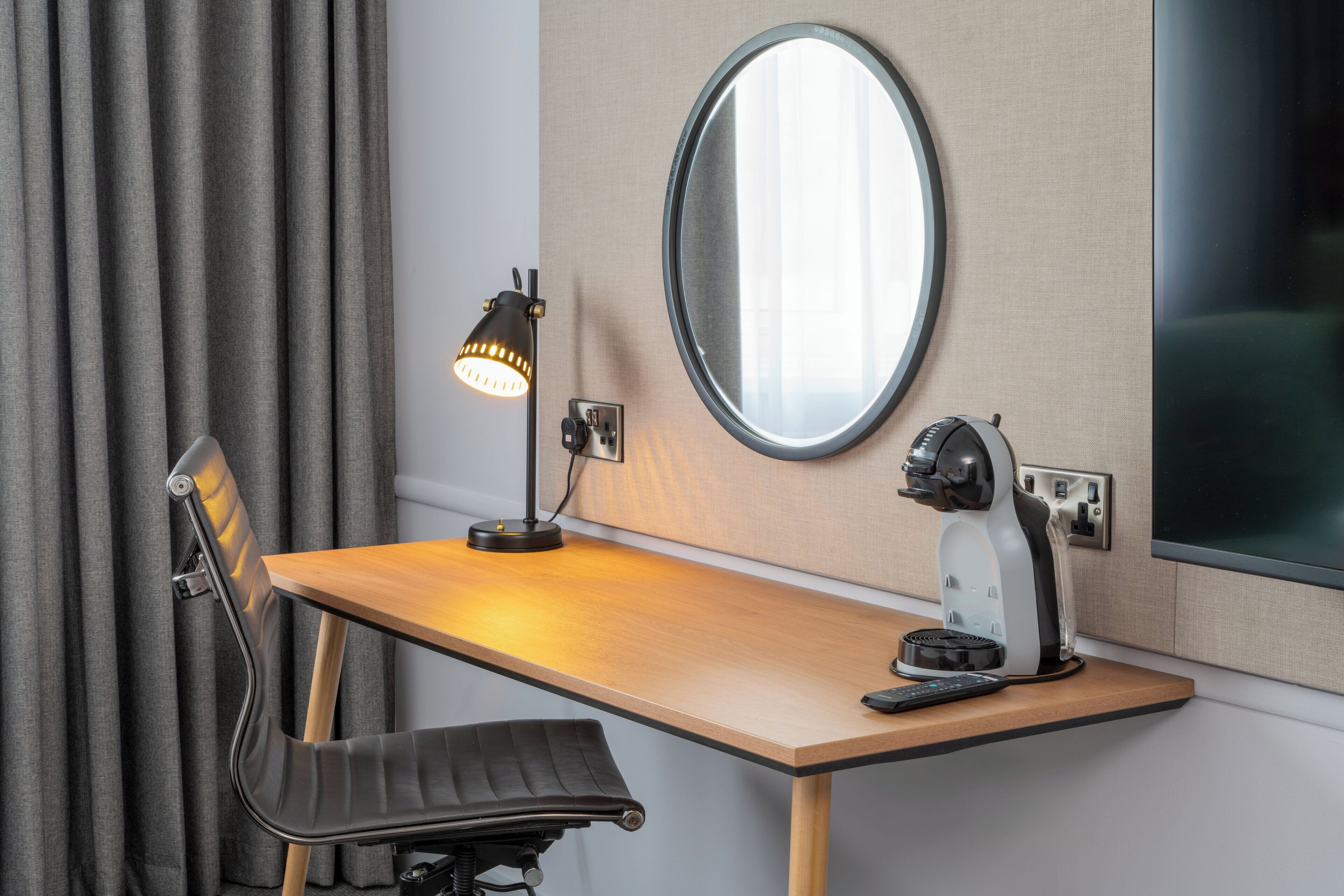 A room with a wooden desk, a black chair, a round mirror, a lamp, and a coffee machine.