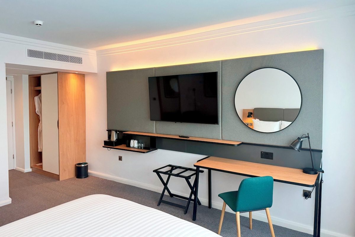 A hotel room with a bed, a desk, a chair, a television, a mirror, and a wardrobe.