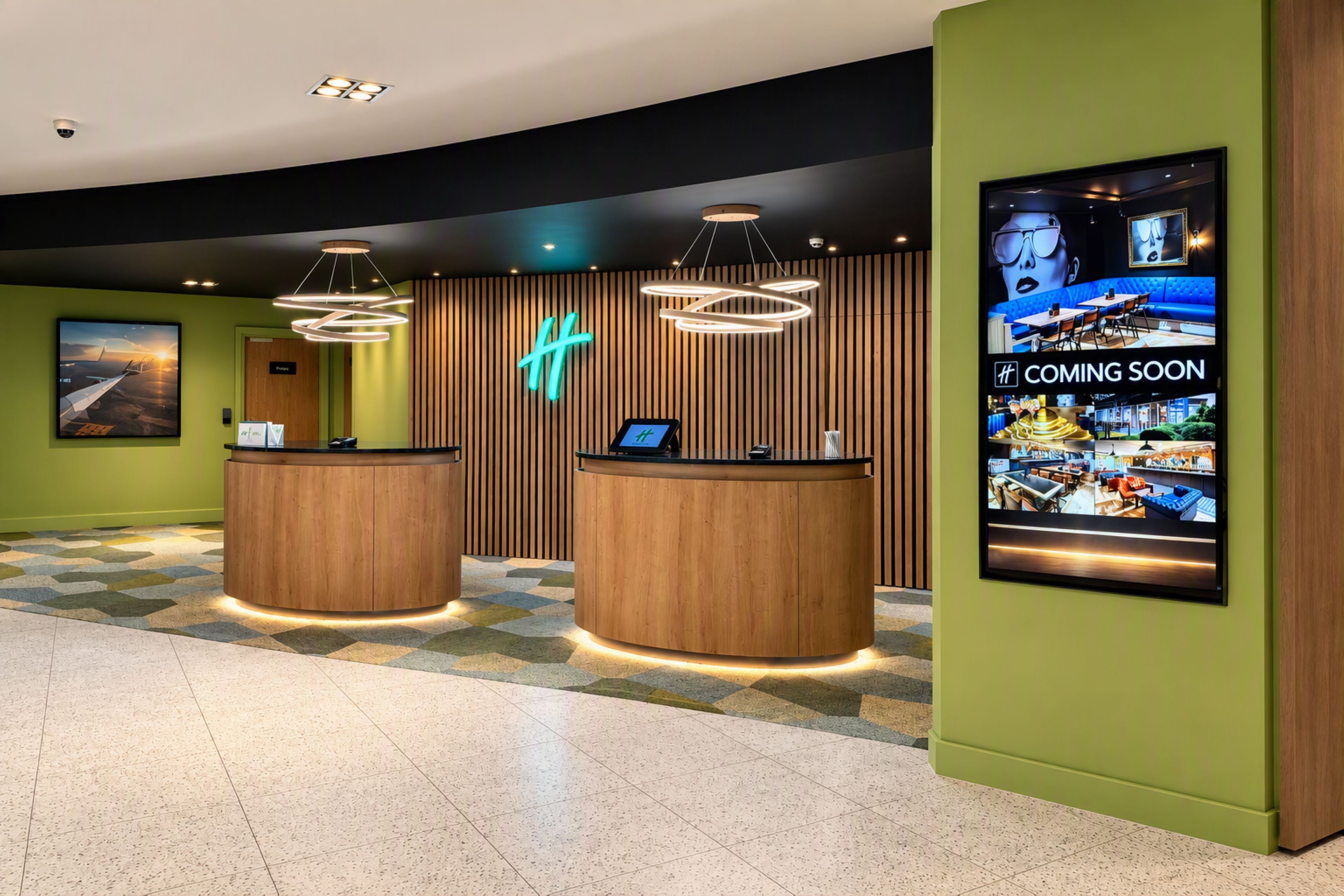 A hotel reception area with two wooden reception desks, a neon sign, and a large digital display advertising a new restaurant.