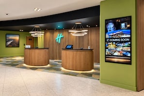 Un hall d'accueil d'hôtel avec deux comptoirs de réception en bois, un panneau lumineux au néon et un grand écran numérique affichant une publicité pour un nouveau restaurant.