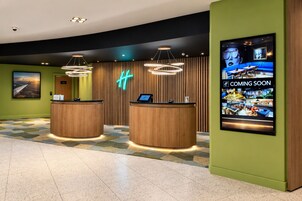 Zona de recepción de un hotel con dos mostradores de madera, un letrero de neón y una gran pantalla digital que anuncia un nuevo restaurante.