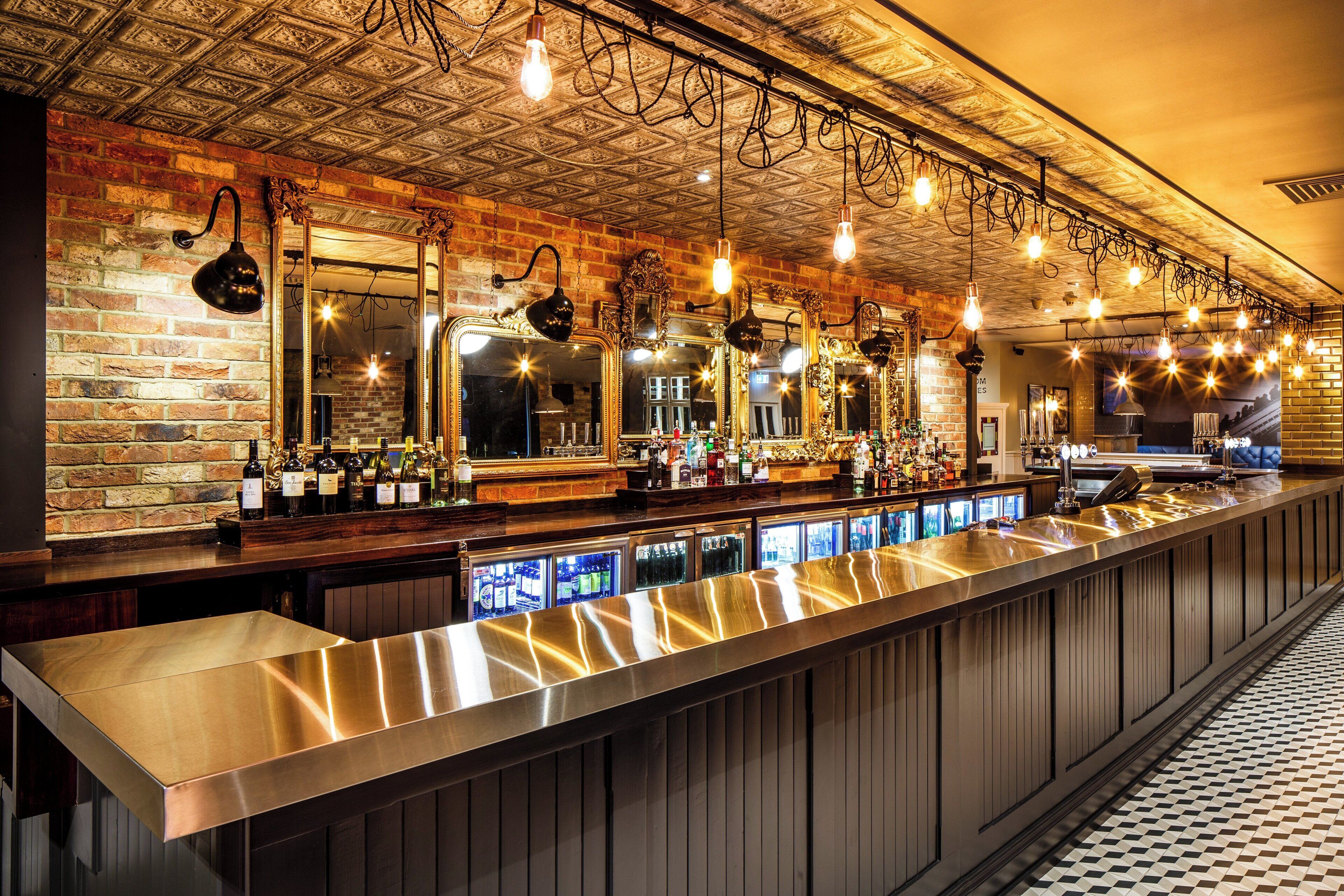 A bar with a long counter, multiple mirrors, hanging light bulbs, and a brick wall.