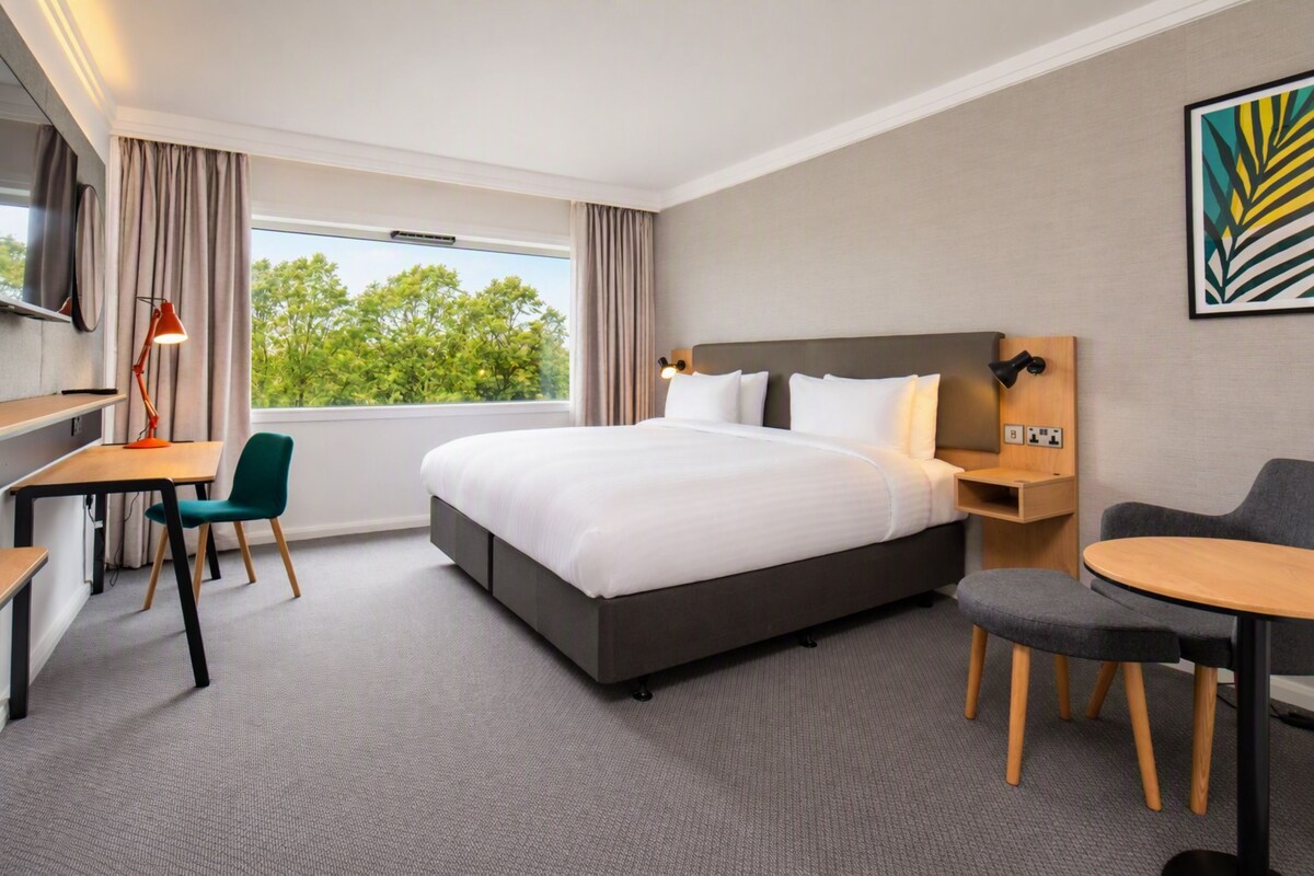 A hotel room with a large bed, a desk, a chair, a small table, and a TV.