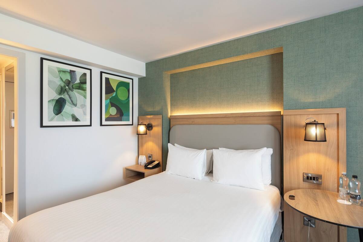 Une chambre d’hôtel avec un grand lit, deux tables de chevet, une petite table et deux lampes fixées au mur.