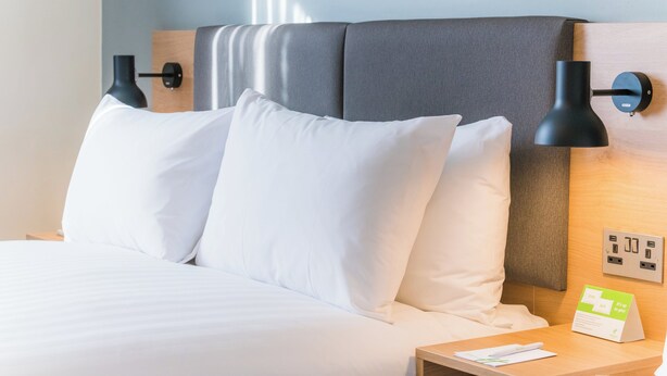 A neatly made bed with white linens, a wooden headboard, two black wall lamps, a bedside table with a lamp, and a sign indicating room amenities. - Holiday Inn London - Heathrow M4,jct.4 by IHG (West Drayton)