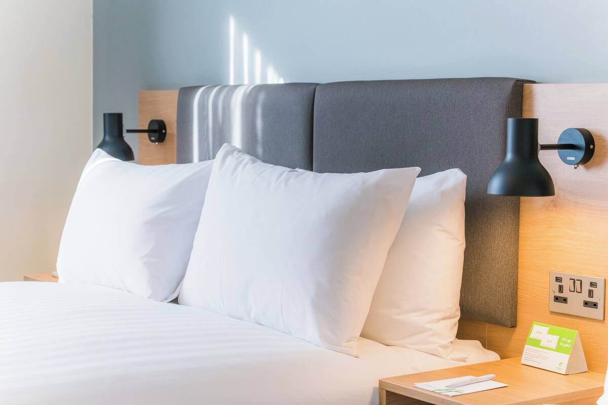 Un lit bien fait, avec du linge de lit blanc, une tête de lit en bois, deux lampes murales noires, une table de chevet avec une lampe et un panneau indiquant les équipements disponibles dans la chambre.
