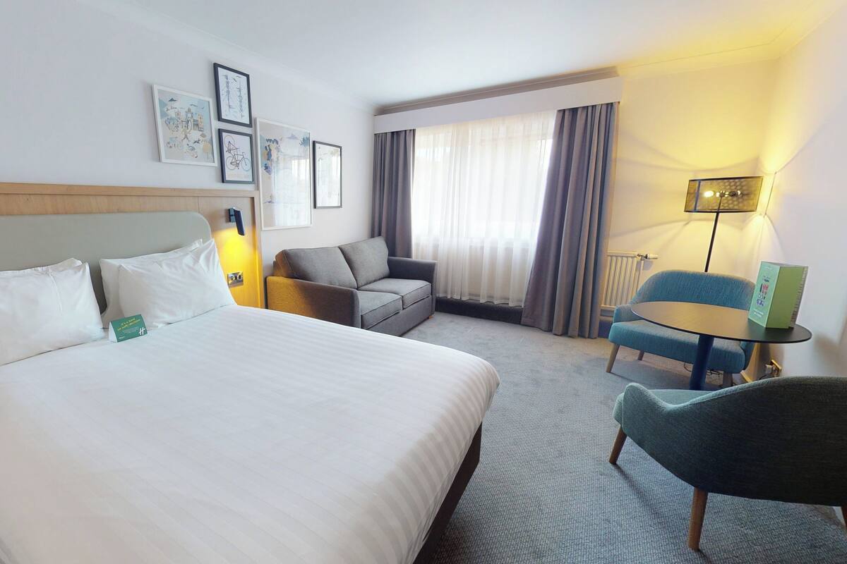 Une chambre d’hôtel avec un grand lit, un canapé, une petite table et une chaise. Des tableaux encadrés ornent le mur, et une lampe se trouve sur la table de chevet.