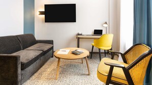 Et moderne hotelværelse med en sofa, en gul stol, et lille rundt bord, et skrivebord med en bærbar computer og et fladskærms-tv på væggen.