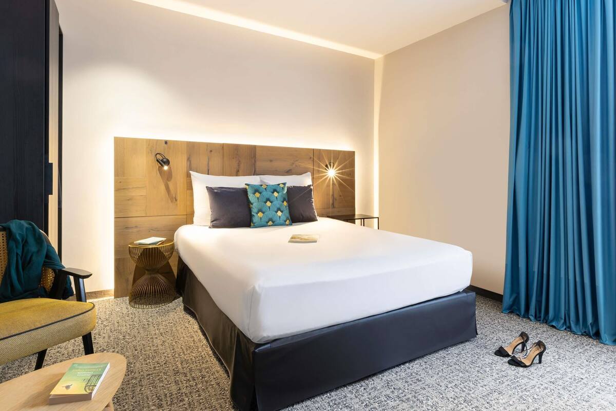 A hotel room with a large bed, a wooden headboard, a bedside table with a lamp, a chair, and a pair of shoes on the floor.