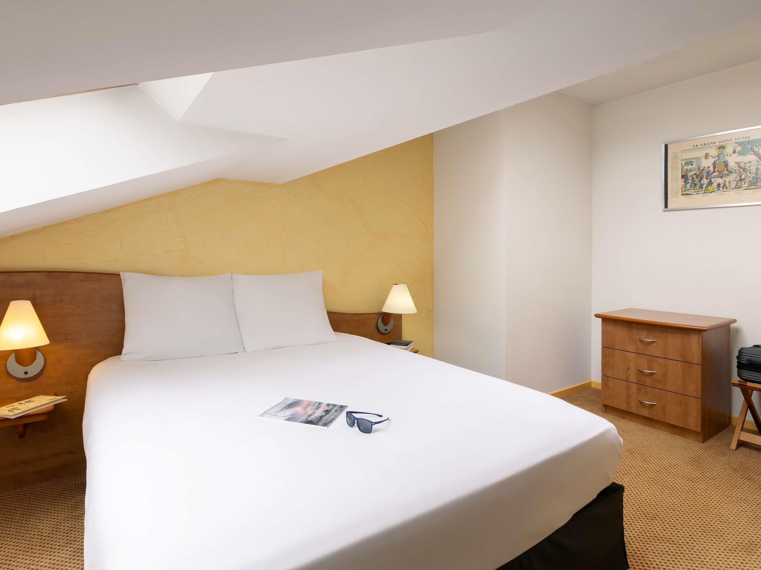 A hotel room with a bed, two bedside lamps, a wooden nightstand, a small wooden dresser, and a framed picture on the wall.