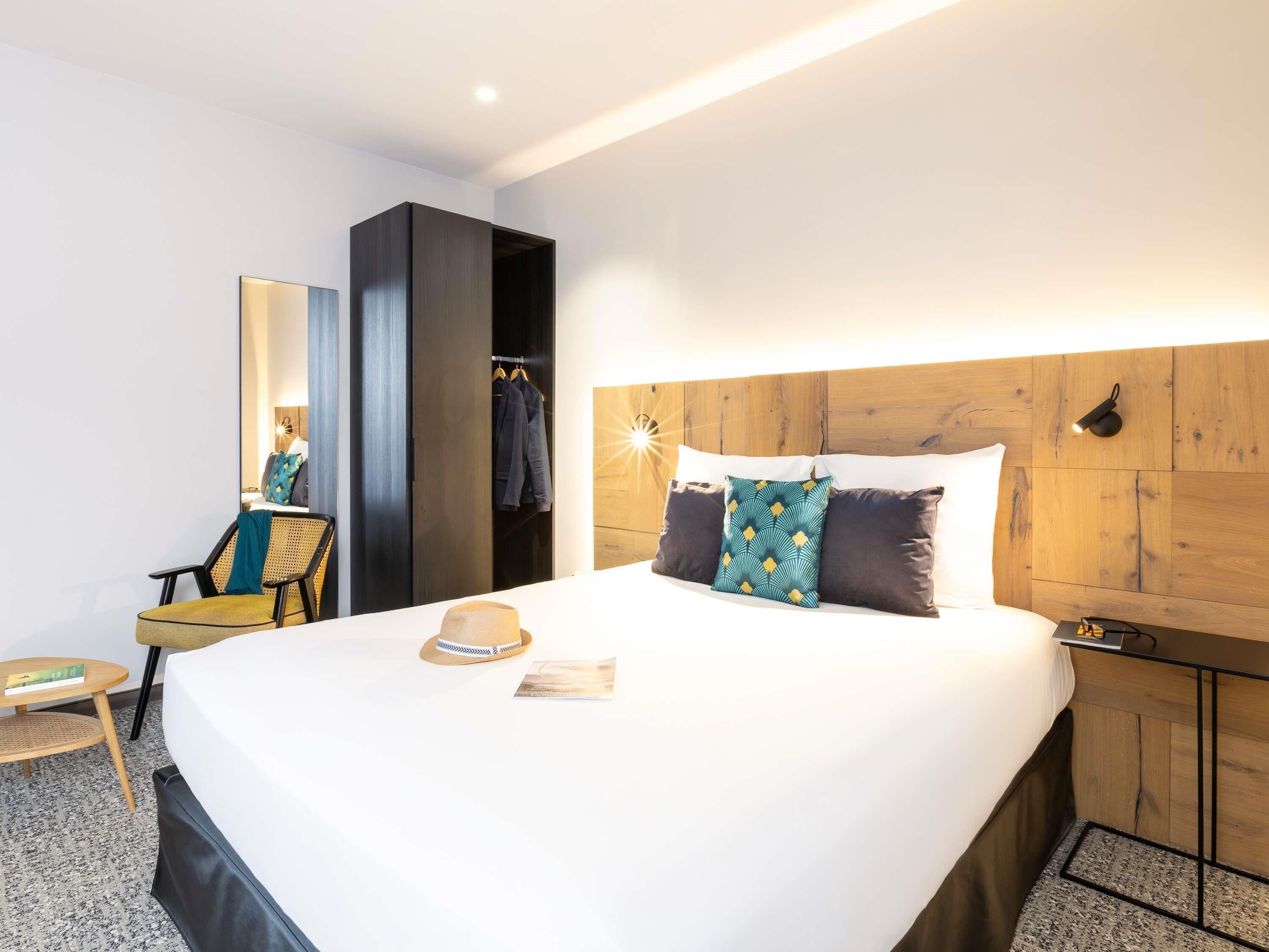 A hotel room with a large bed, a wooden headboard, a wardrobe, a chair, and a small table with a hat and a book.