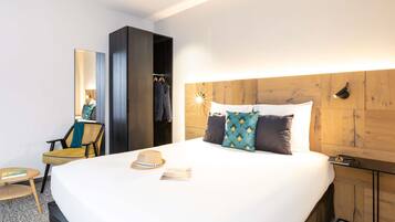 A hotel room with a large bed, a wooden headboard, a wardrobe, a chair, and a small table with a hat and a book.