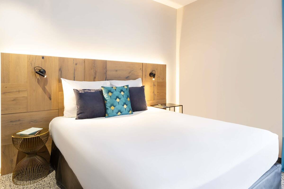 A neatly made bed with a wooden headboard, two bedside lamps, and a small round table with a book and glasses.