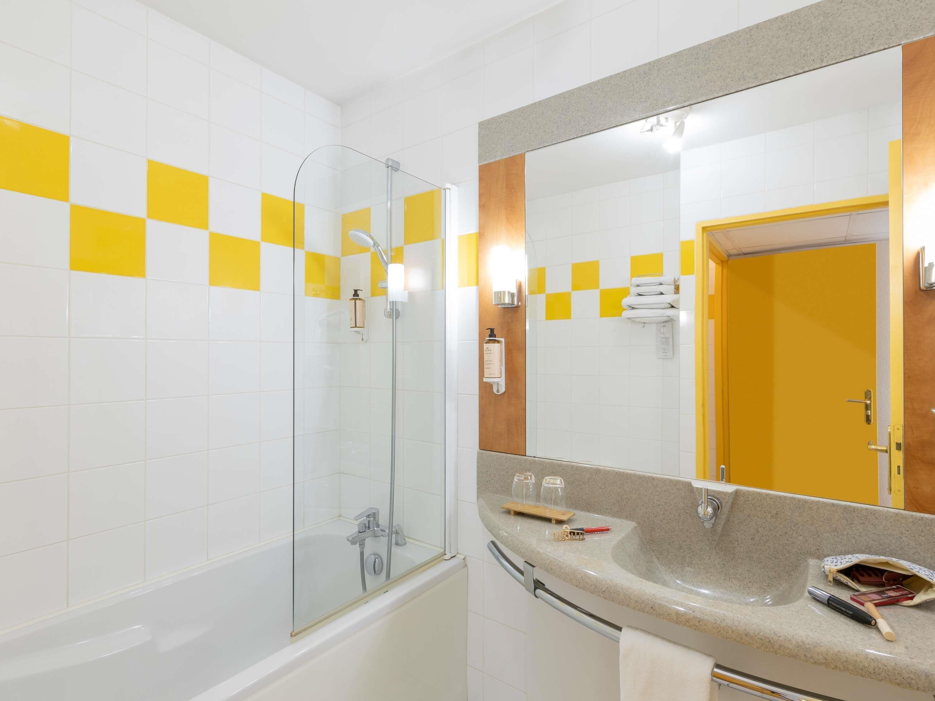 A bathroom with a glass-enclosed shower, a sink with toiletries, and a mirror reflecting a yellow door.