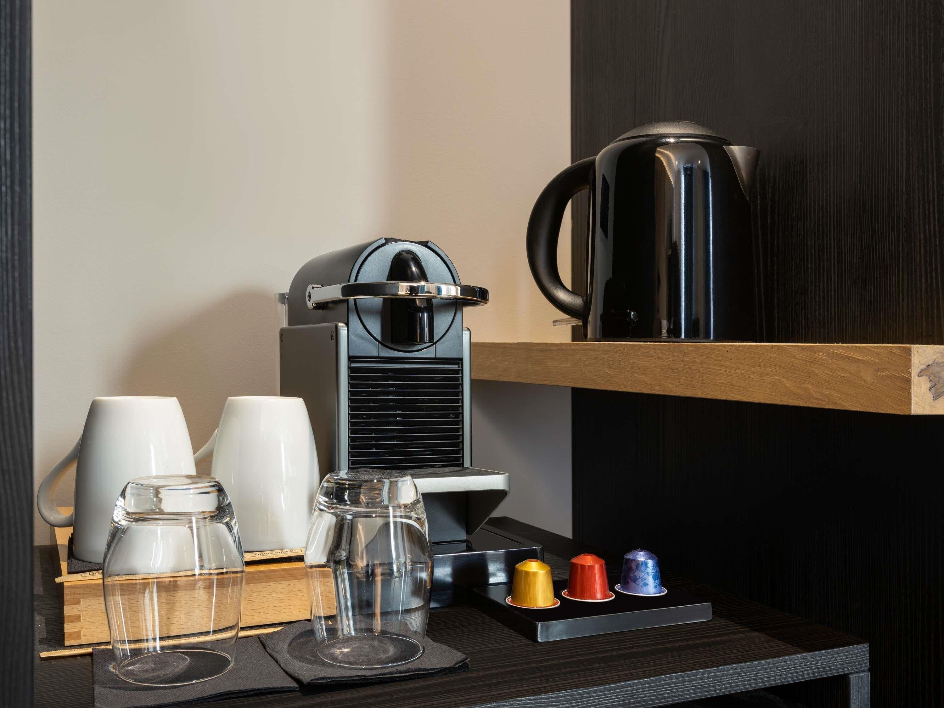 A coffee machine, two white cups, a glass container, and colorful lids on a table.