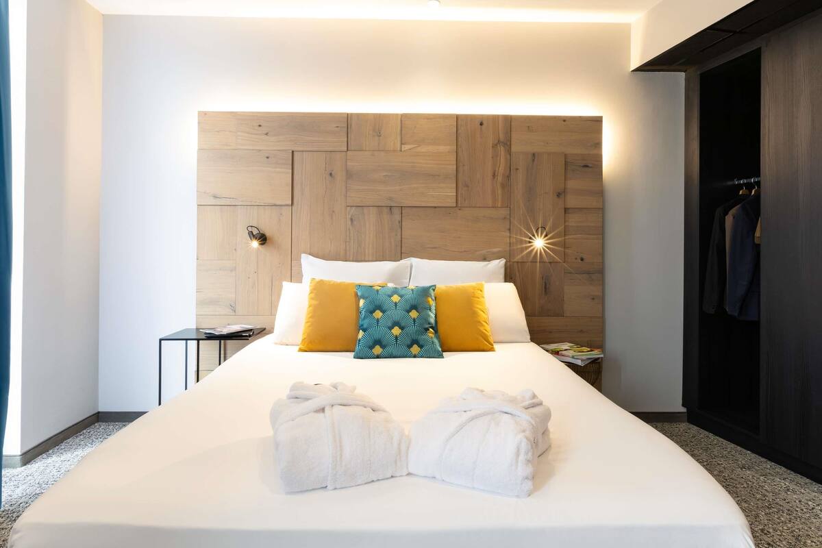 A modern hotel room with a wooden headboard, white bedding, and a pair of towels on the bed.