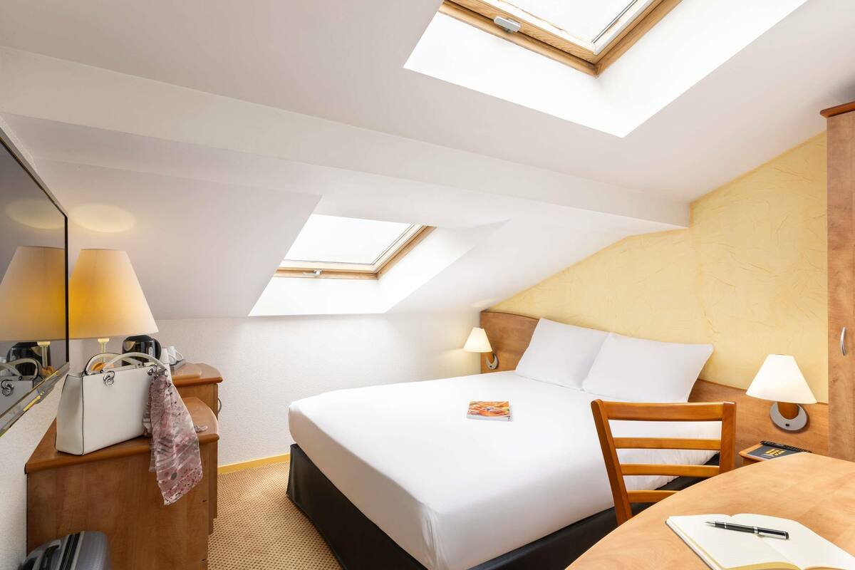 A hotel room with a bed, a desk, a chair, a suitcase, a lamp, and a mirror.