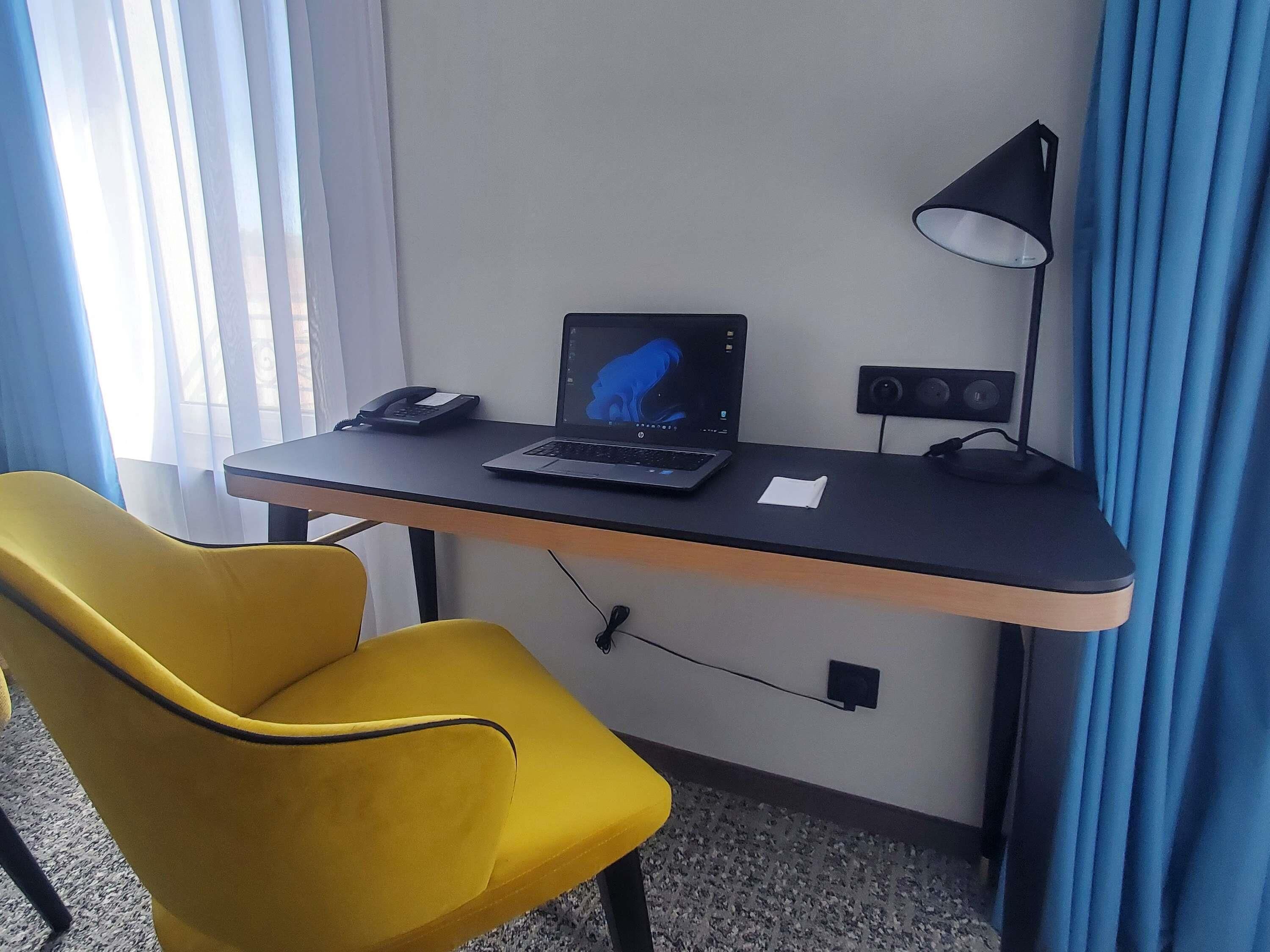 A compact hotel room with a desk, a laptop, a telephone, a lamp, and a yellow chair.