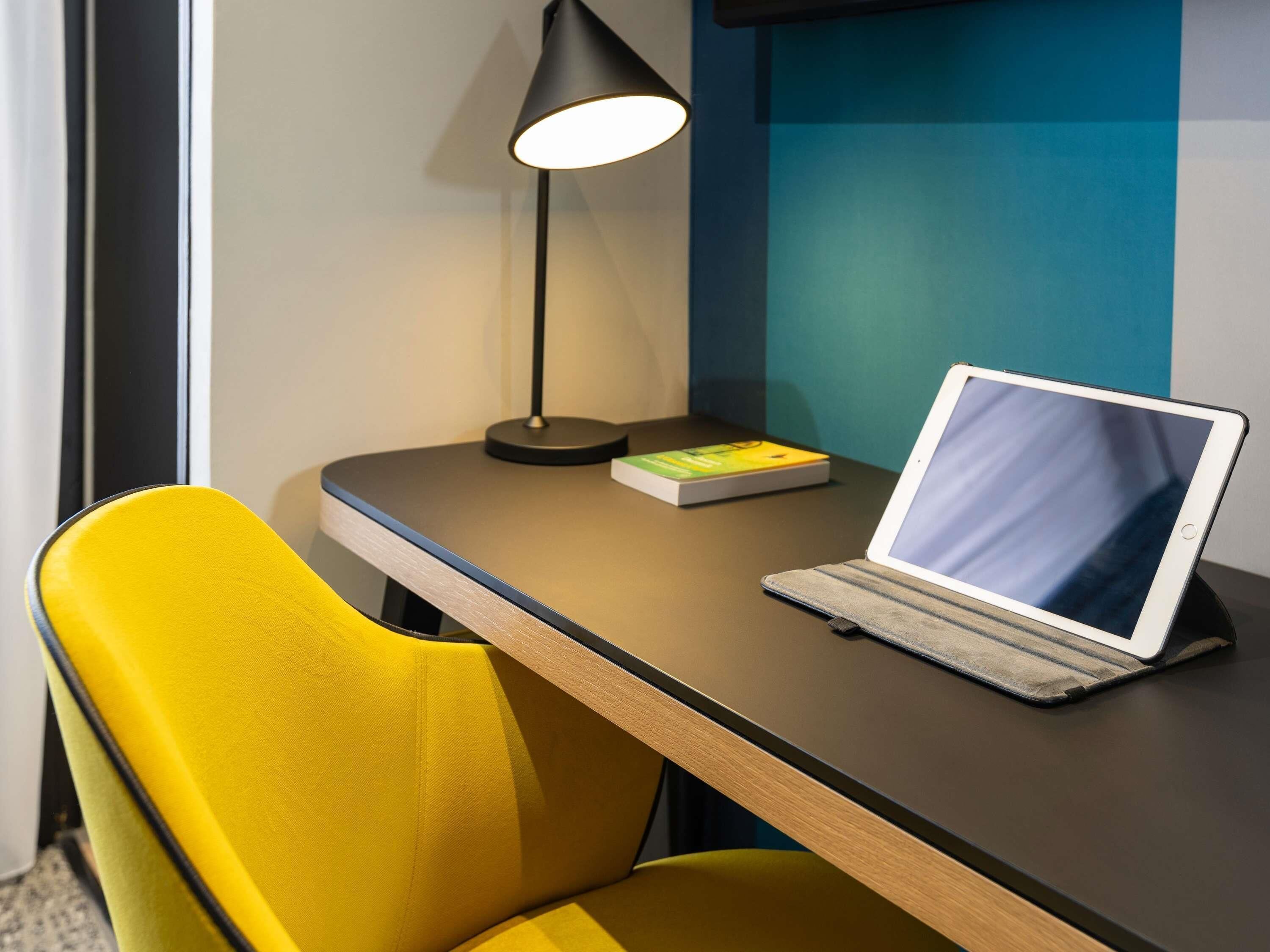 A laptop on a desk with a book, a lamp, and a yellow chair.