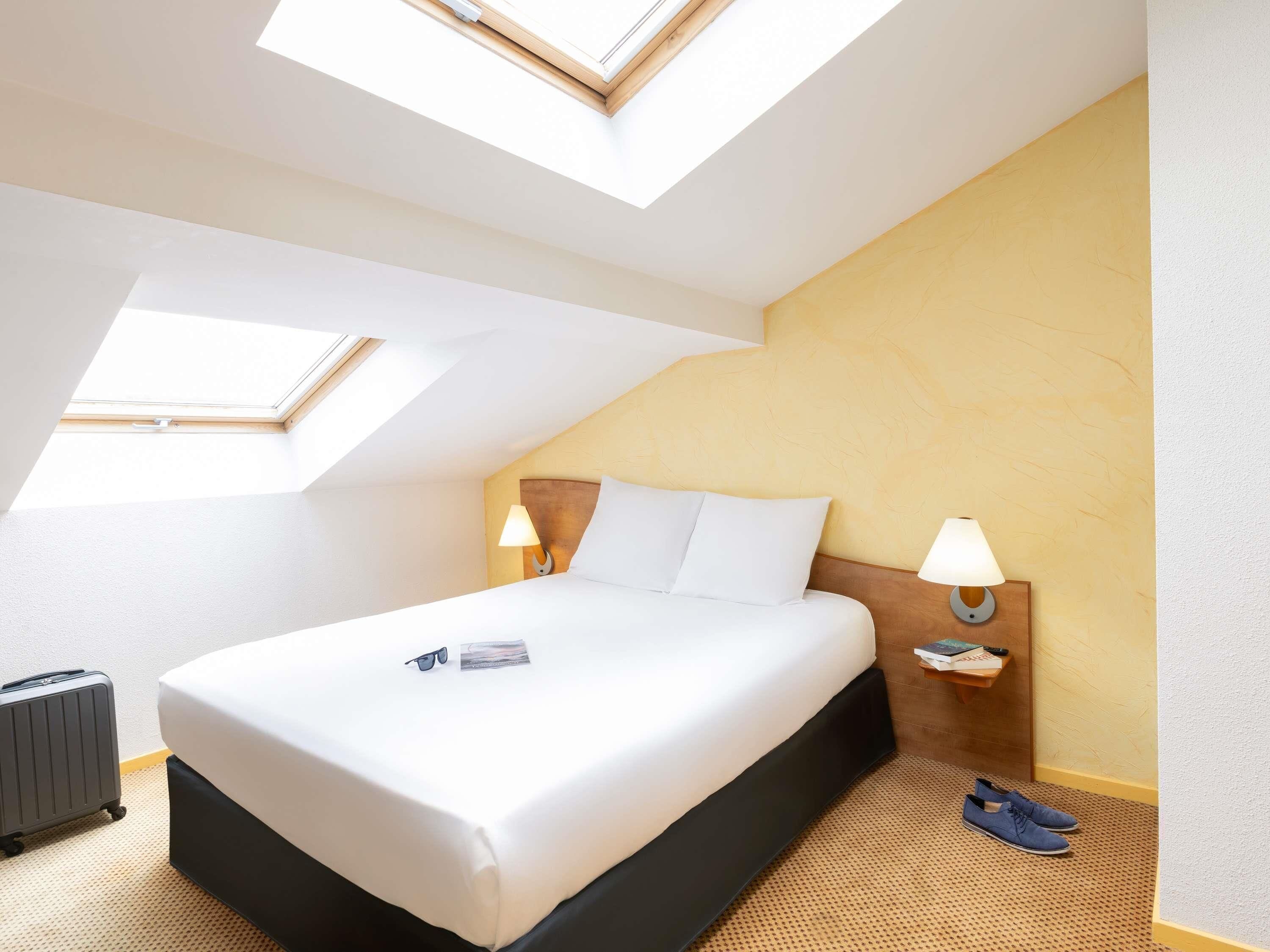 Habitación de hotel con cama, dos lámparas de noche, una mesita pequeña, un par de zapatos y una maleta.