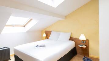 Habitación de hotel con cama, dos lámparas de noche, una mesita pequeña, un par de zapatos y una maleta.