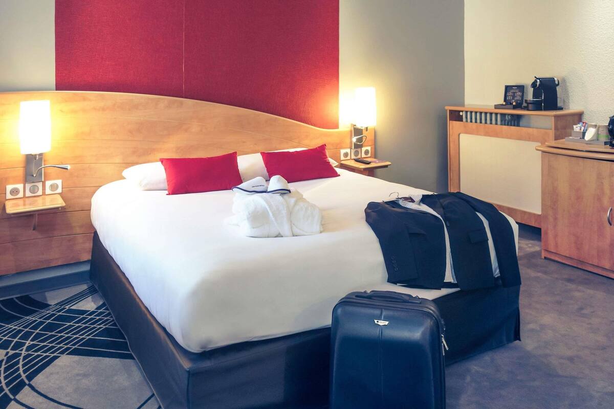 A hotel room with a bed, two red pillows, a white bathrobe, a black suitcase, and a wooden headboard.