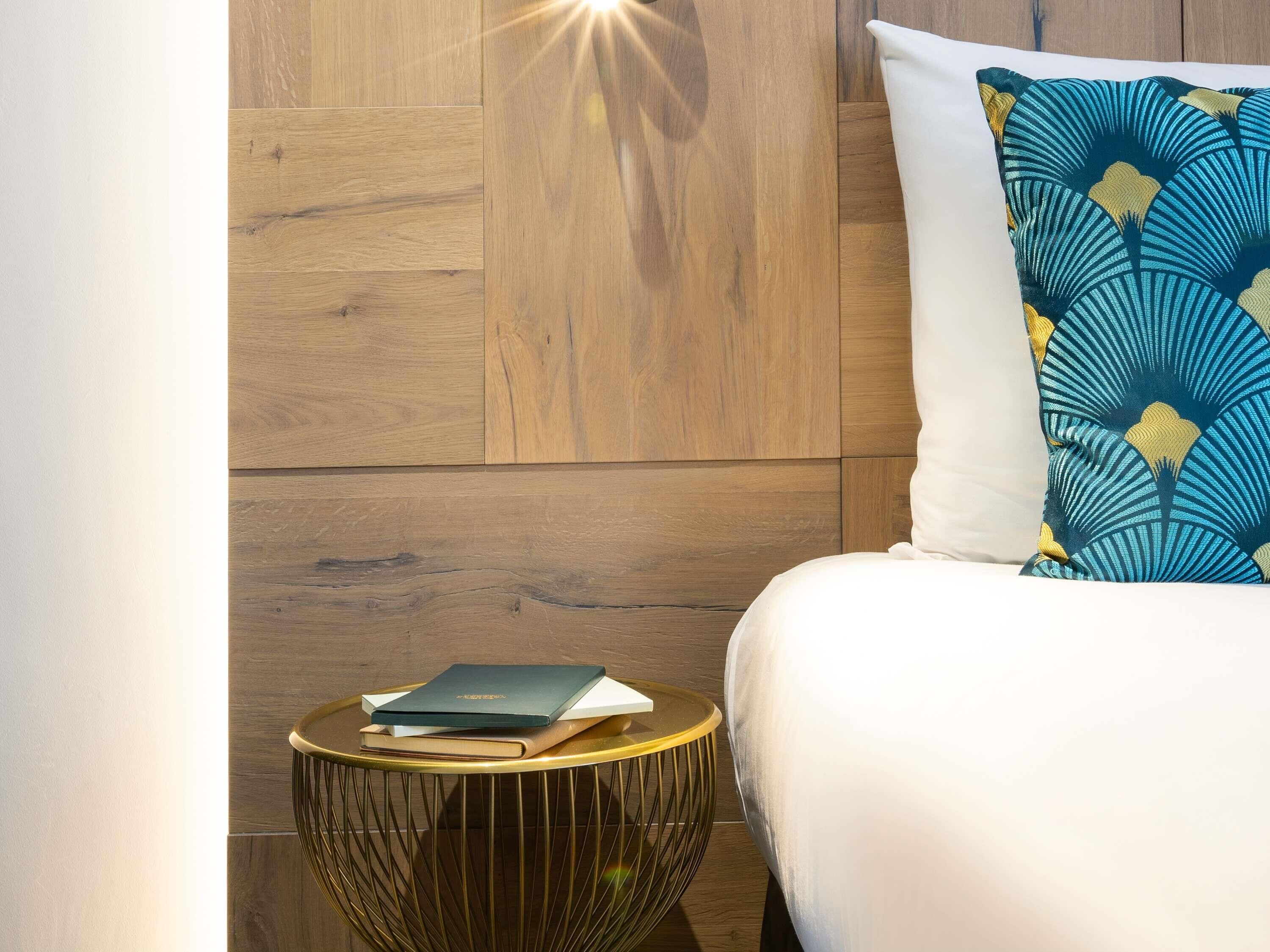 A bedroom with a wooden wall panel, a white bed, a patterned pillow, and a small round side table with a book.