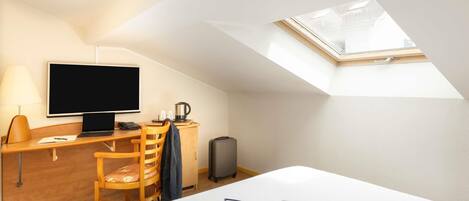 A hotel room with a desk, chair, bed, TV, and a skylight.