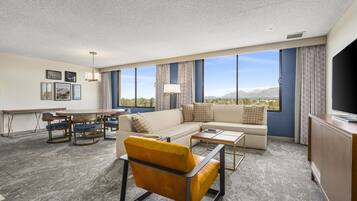 A hotel room with a sofa, armchair, dining table, and a large window offering a mountain view.