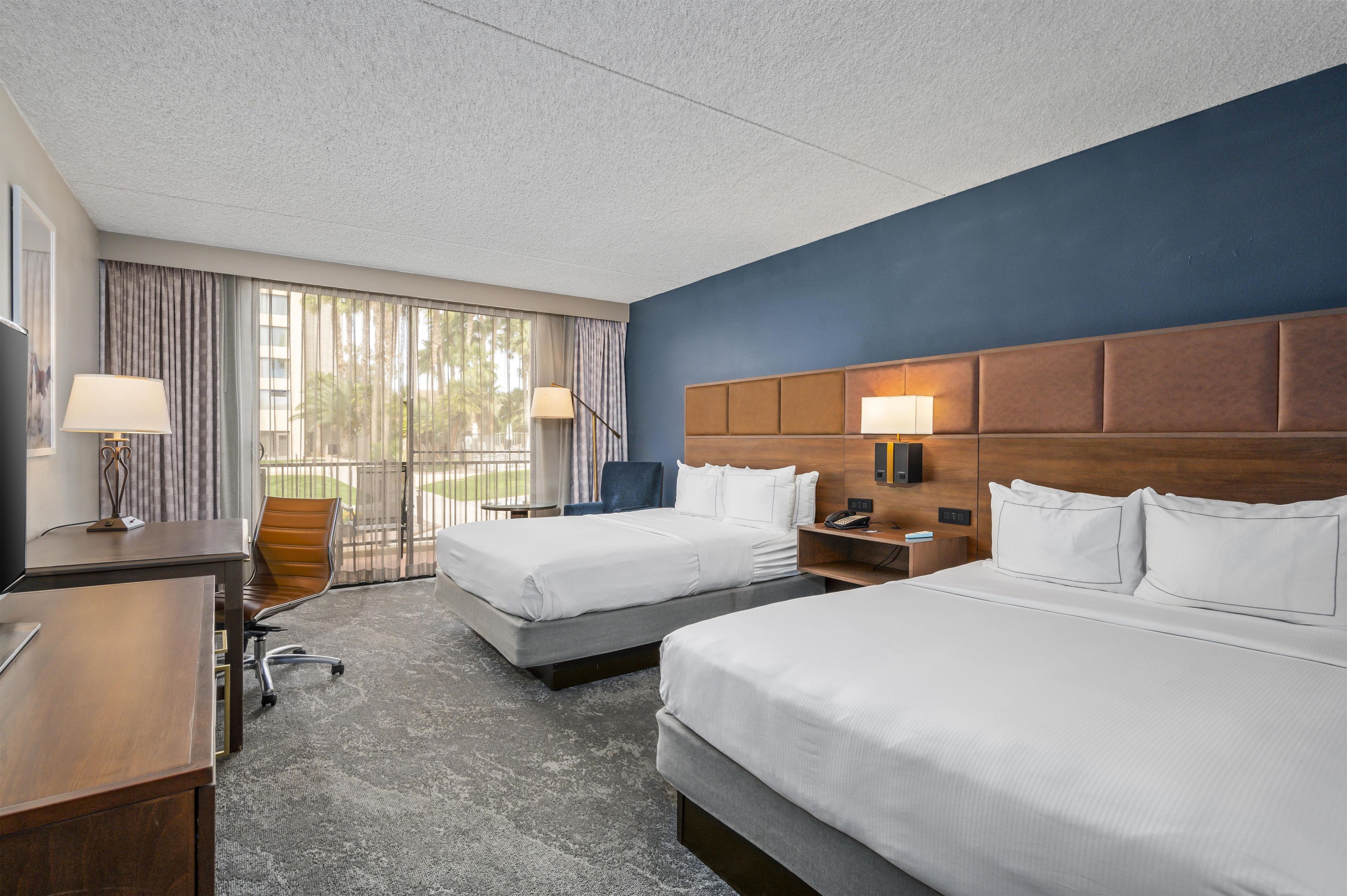A hotel room with two beds, a desk, a chair, and a view of palm trees through the window.