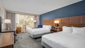 A hotel room with two beds, a desk, a chair, and a view of palm trees through the window.