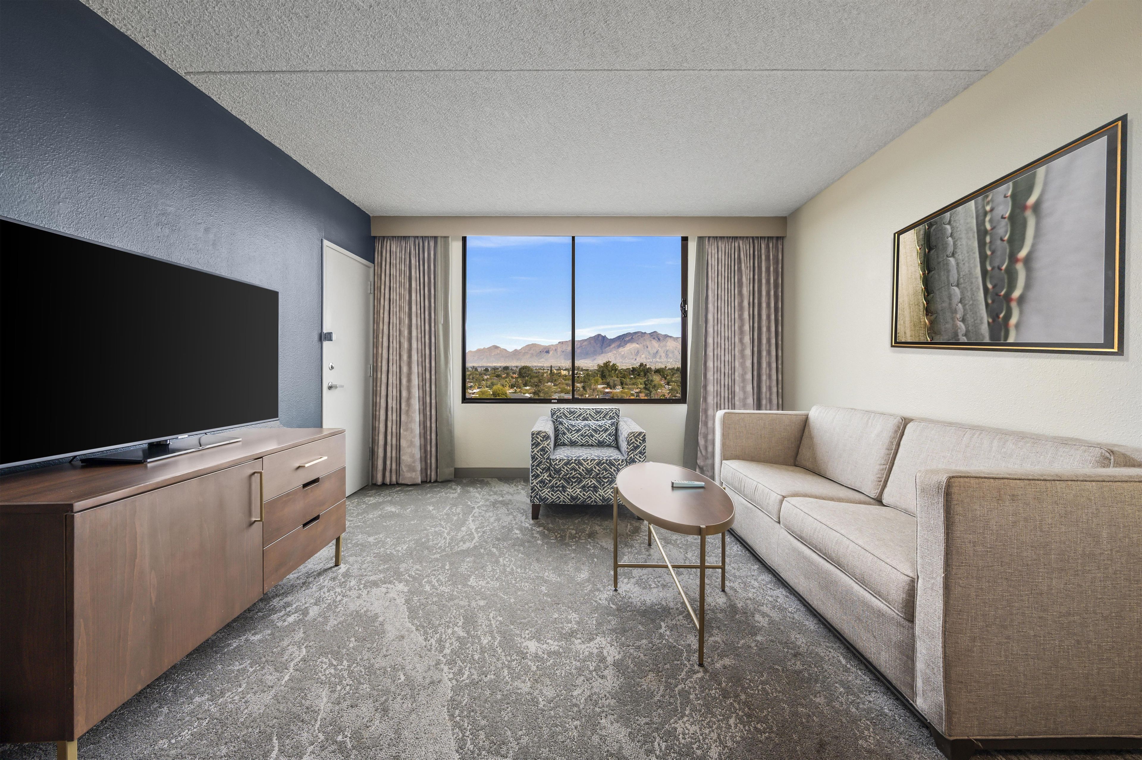 A hotel room with a large flat-screen TV, a sofa, a patterned armchair, and a small table.