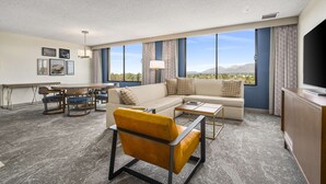 Una habitación de hotel moderna con sofá, sillón, mesa de comedor y un amplio ventanal con vistas a la montaña.