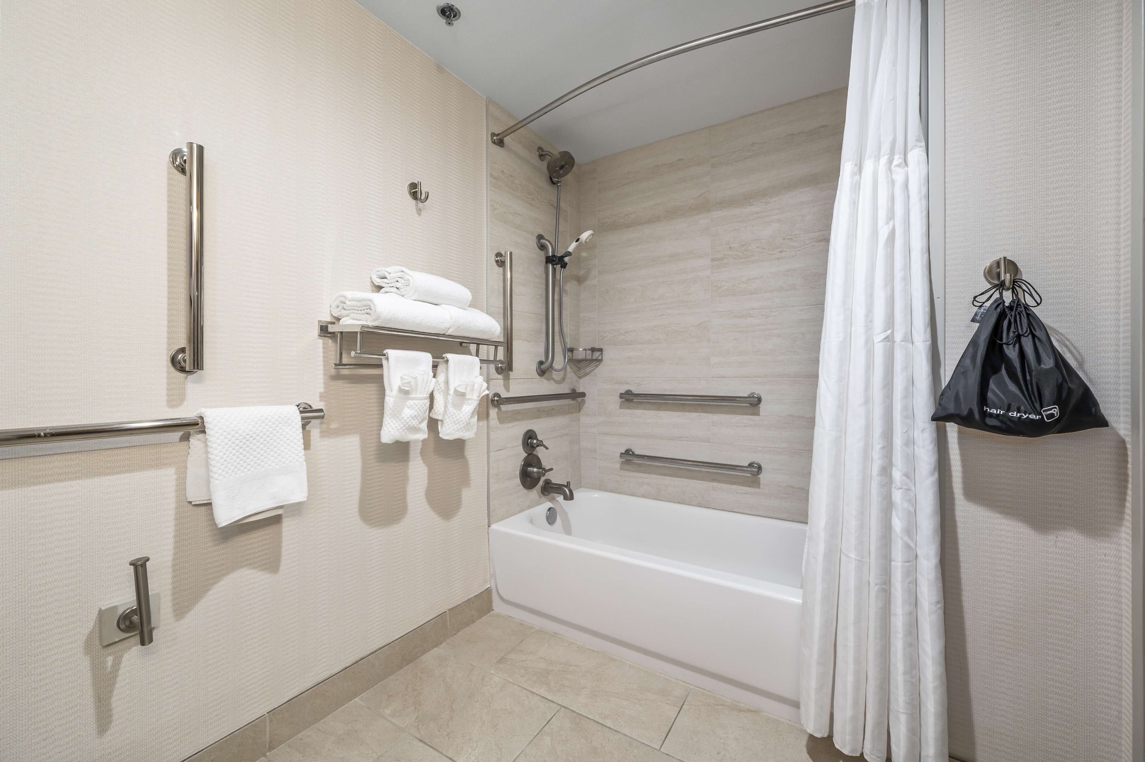 A modern bathroom with a bathtub, a shower, and a handrail. There are towels on a shelf and a hair dryer hanging on the wall.