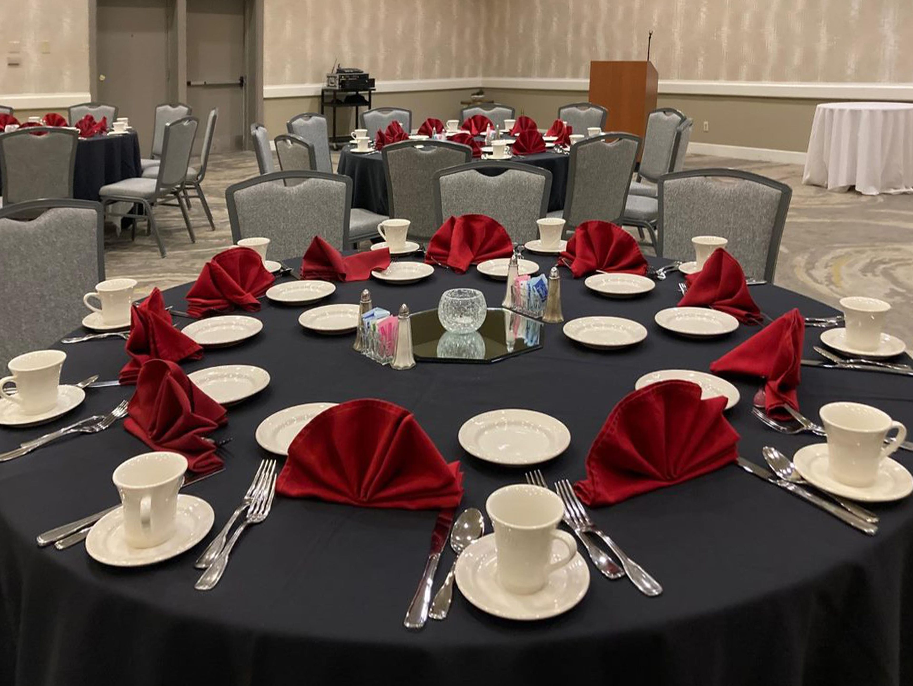 Une salle de conférence avec des tables rondes dressées pour un événement formel, agrémentée d’assiettes blanches, de couverts en argent et de serviettes rouges.