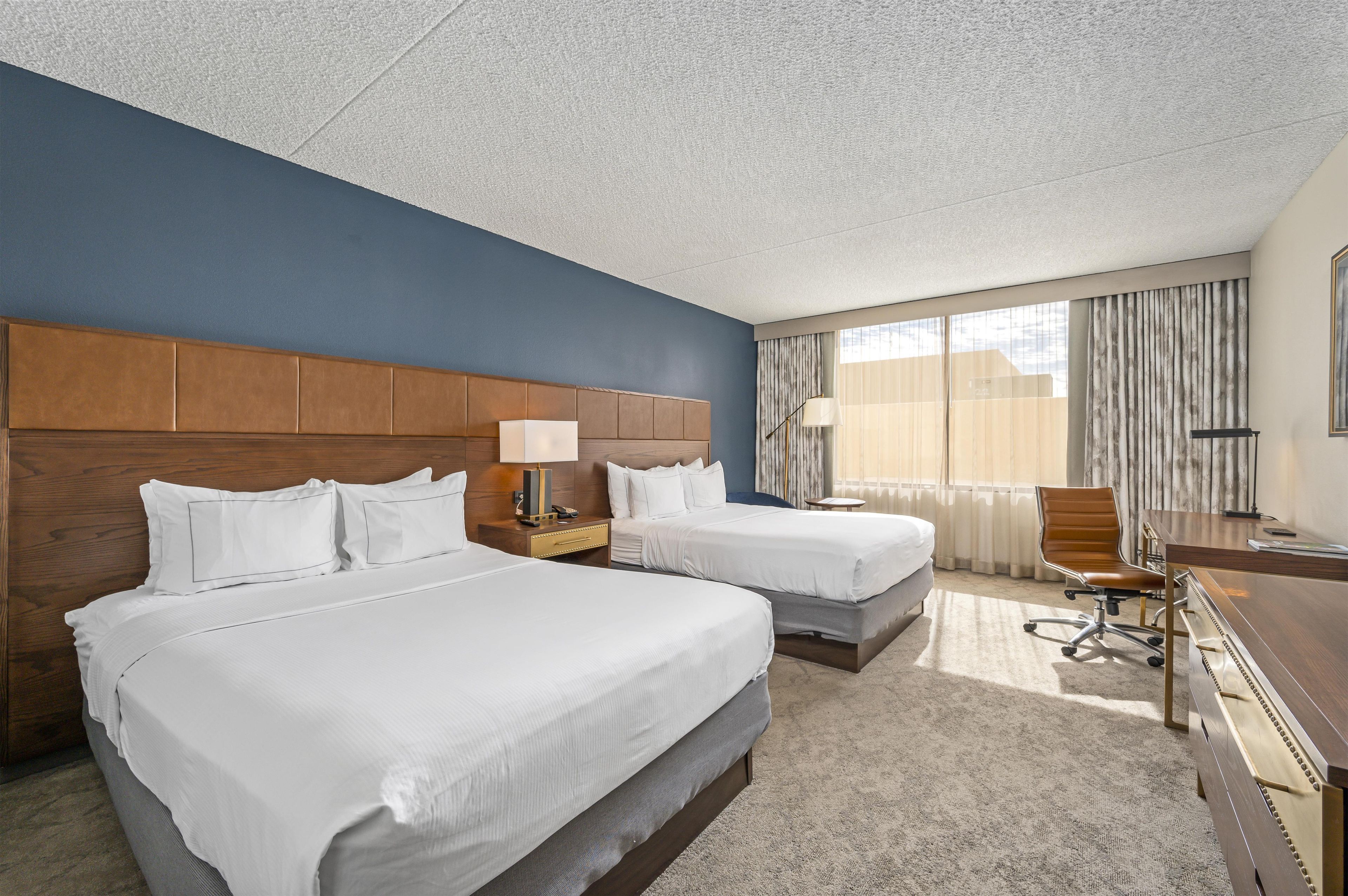 A hotel room with two beds, a desk, a chair, and a large window with curtains.