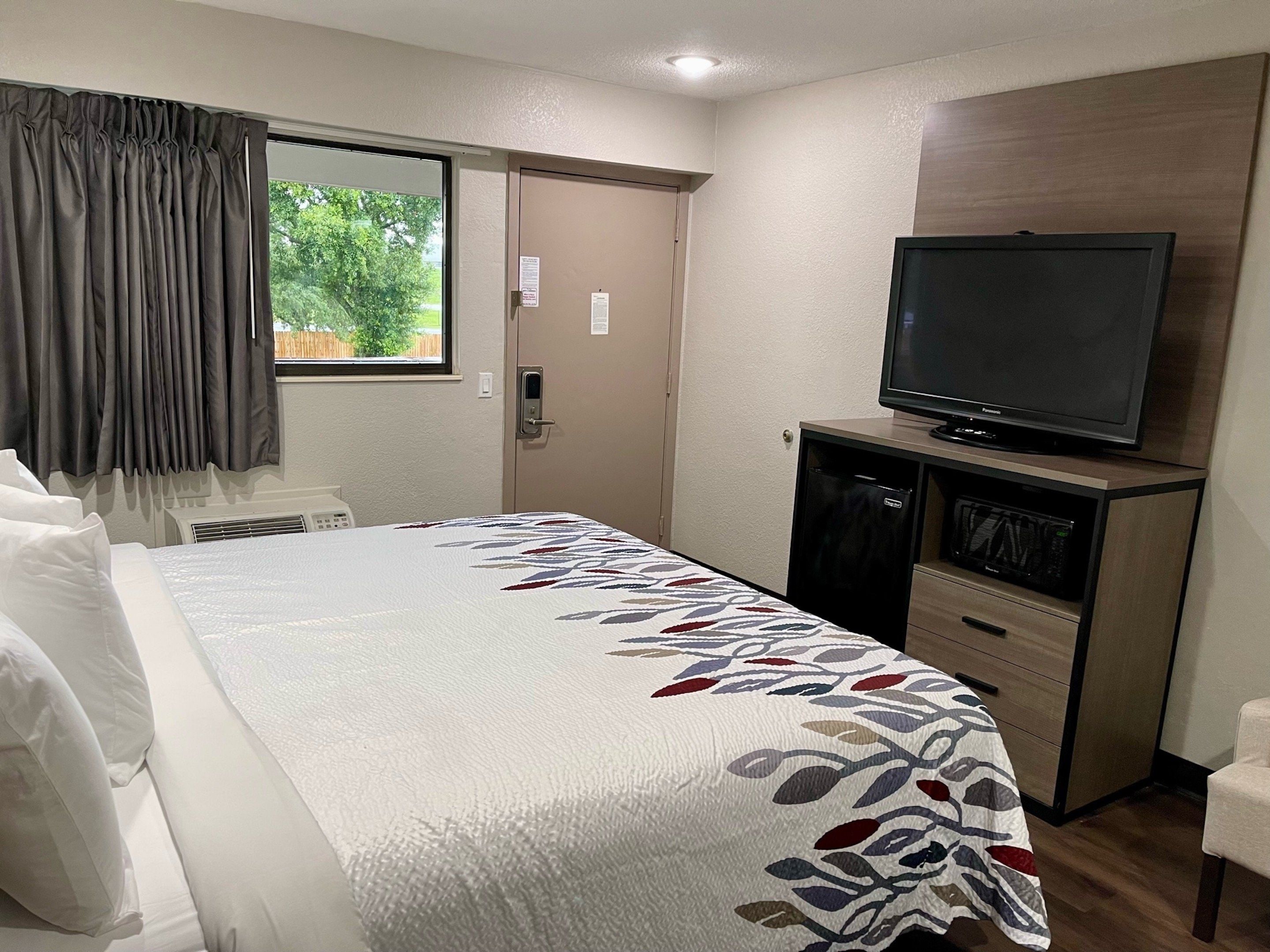 Ett hotellrum med en säng, en tv på ett stativ, ett skrivbord med en dator och ett fönster med gardiner.