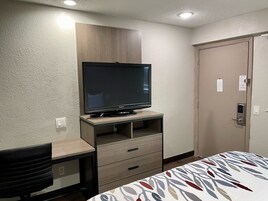 Quarto de hotel com televisão sobre suporte de madeira, cama com colcha estampada, escrivaninha com cadeira e porta com fechadura.