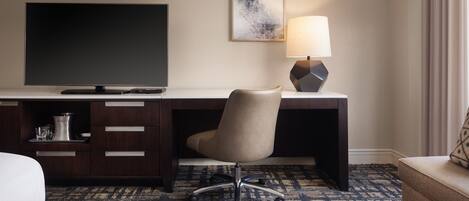 A hotel room with a dark wood desk, a flat-screen TV, a modern desk lamp, a cushioned office chair, a patterned carpet, and a bed with a white comforter.