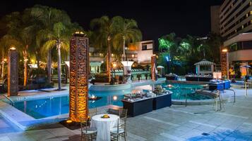 A nighttime pool area with multiple pools, palm trees, and a dining setup.